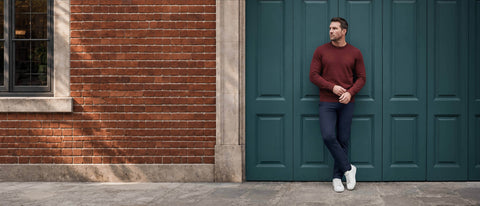A man stands against a dark teal paneled door next to a red brick wall, wearing a burgundy knit sweater, dark blue jeans, and white sneakers.