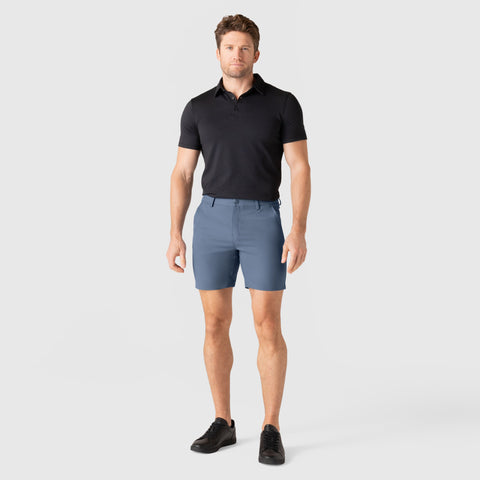 Man wearing Jetsetter Tech Short in blue, paired with a black polo shirt and black shoes.