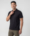 A man stands against a plain light background, smiling and wearing the Jack Archer Clubhouse Pique Polo in True Black and olive green pants, holding the collar of his moisture-managing shirt with one hand.