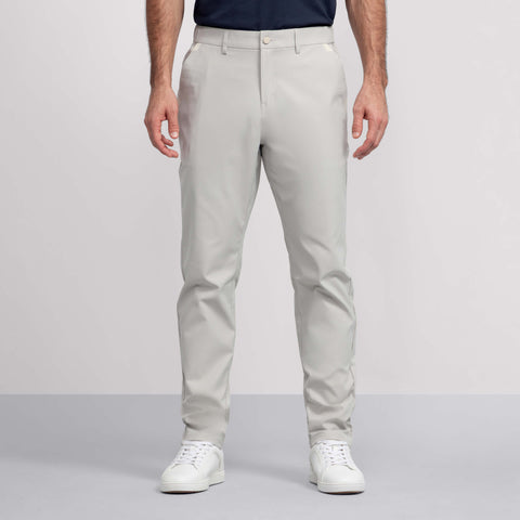 A man wears Jack Archer Jetsetter Tech Pant Relaxed Fit in Sand Stone with a dark tucked-in shirt and white sneakers, standing with arms at his sides against a plain background. Only his upper body is partially visible.