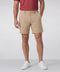 A man stands against a plain light background wearing a red short-sleeve shirt, white sneakers, and Jack Archer’s Jetsetter Tech Short 7" in Sand Tan, with one hand in his pocket.