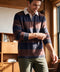 A brown and navy striped rugby polo with a white collar, worn with olive chinos indoors.