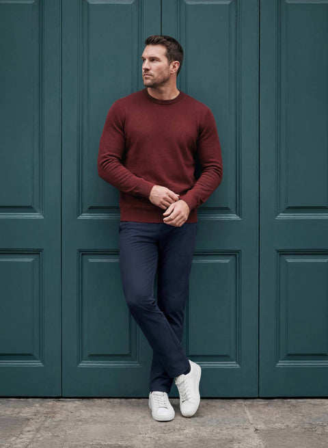 A man wearing a burgundy long-sleeve crewneck sweater, navy blue trousers, and white sneakers leans against a dark teal paneled wooden door.
