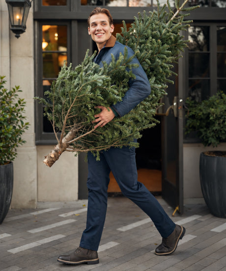 A man in a navy quilted jacket and navy pants carrying a Christmas tree past a storefront.