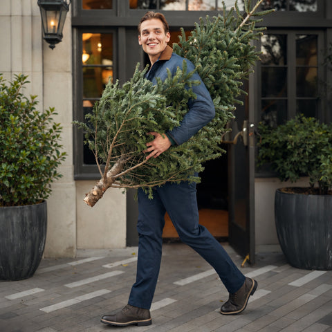 A man in a navy quilted jacket and navy pants carrying a Christmas tree past a storefront.
