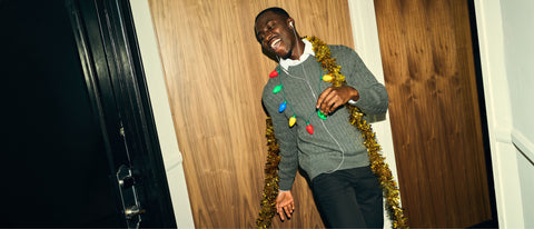 A man in a grey cable-knit sweater draped in gold tinsel and a string of holiday lights.