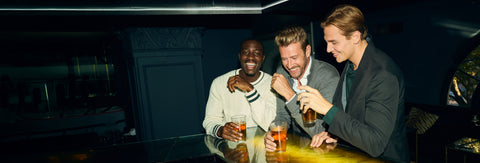 Three friends in cream knitwear and blazers share drinks at a dimly lit bar.