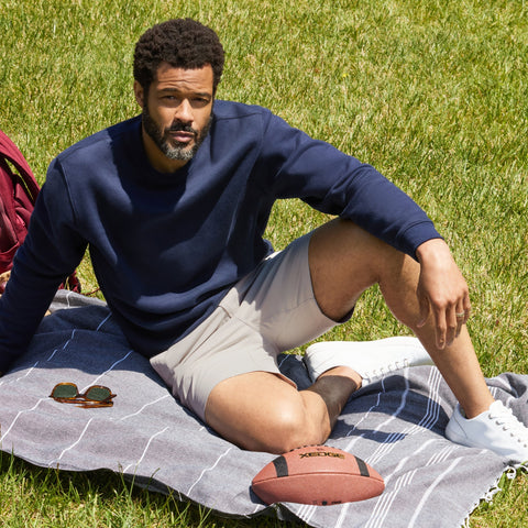 Man wearing Golf Game Essentials shorts and navy sweater, sitting on a picnic blanket outdoors.