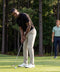 On a tree-lined golf course, one man putts in Jack Archer's Clubhouse Pique Polo in True Black and beige pants, while another, dressed in a light green four-way stretch polo and navy pants, watches from behind holding a club.