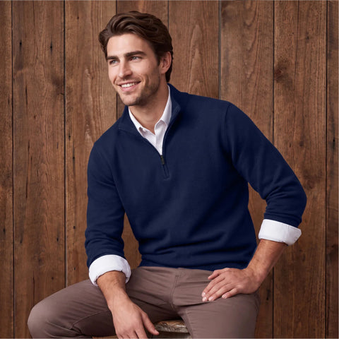 A man in a navy quarter-zip sweater over a white shirt and taupe pants, seated on a stool.