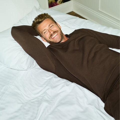 A man in a brown sweatshirt and matching sweatpants stretched out on a white bed.