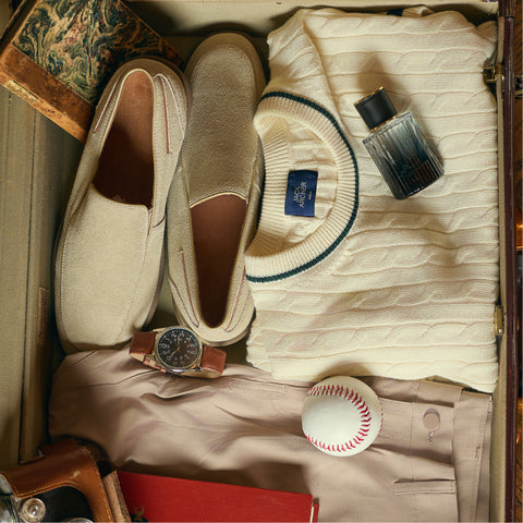 A suitcase flat-lay with suede loafers, a cream cable-knit sweater, cologne, a watch and a baseball.