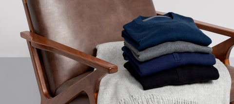 Four folded sweaters in navy, grey and black stacked on a blanket over a leather lounge chair.