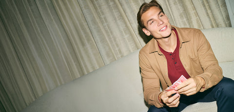 A man in a camel overshirt over a red henley holding playing cards on a cream sofa.