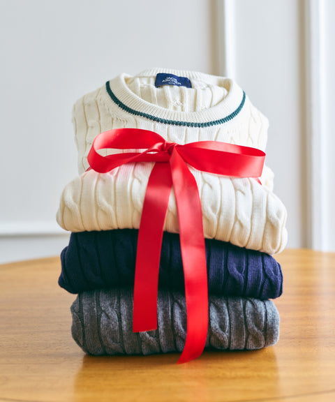 A stack of cream, navy and grey cable-knit sweaters tied with a red gift ribbon.