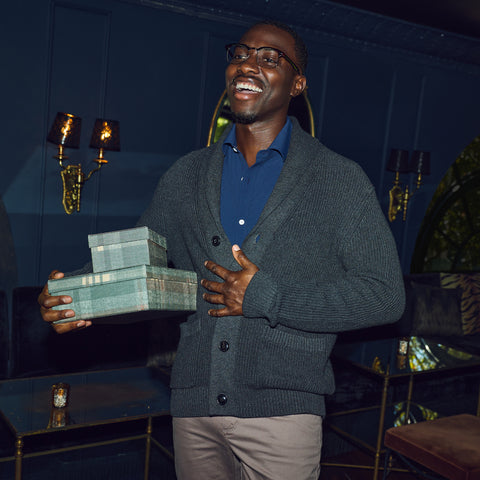 A man in a charcoal shawl-collar cardigan and blue polo holding a stack of wrapped gift boxes.