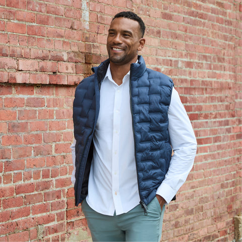 A man in a navy puffer vest over a white shirt and sage pants against a brick wall.