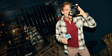 A partygoer in a cream plaid overshirt and maroon waffle henley takes a Polaroid at a dim bar.