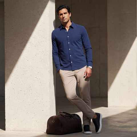 Taupe Jetsetter pants and a navy button-down shirt, worn beside a leather duffel against a concrete wall.
