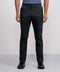 Wearing Jack Archer's Jetsetter Tech Pant—a dark, water-repellent, wrinkle-resistant pair—plus a blue short-sleeve shirt and black sneakers, a person stands against a plain light gray background, shown from shoulders to feet.