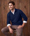 A man in a navy quarter-zip sweater and taupe pants seated against a wood plank wall.