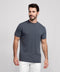 A man with short dark hair and a beard stands against a plain light gray background, wearing the Jack Archer Jetsetter Tee in Charcoal Gray and white pants. He looks slightly left and smiles softly.