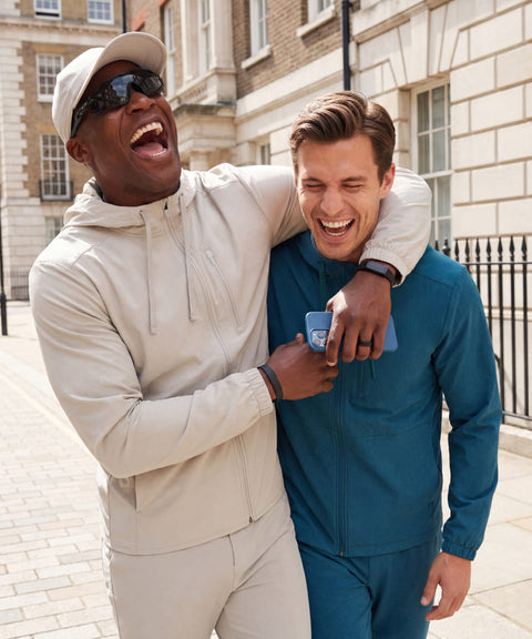 Two men laughing on a city sidewalk in Vapor Gray hooded jacket and Pacific Blue Clubhouse Range set