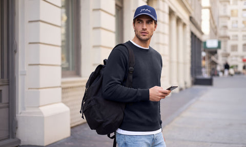 A man in a charcoal crewneck sweatshirt and navy Jack Archer cap walking a city sidewalk with a backpack.