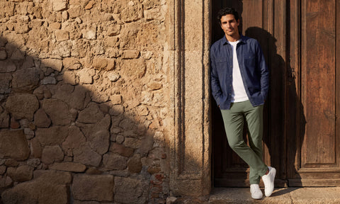 An indigo chambray shirt worn open over a white tee with sage chinos, beside a sunlit stone wall.