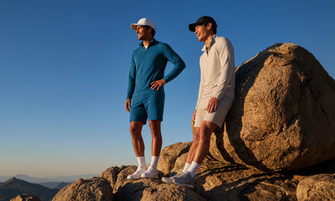 Two men in Clubhouse Range quarter-zips and shorts standing on coastal boulders at sunset