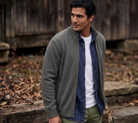 A man in a gray full-zip sweater over a denim shirt, standing against weathered wood siding.