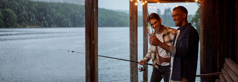 Two men fishing from a covered dock at dusk, one in a plaid overshirt and one in dark denim.