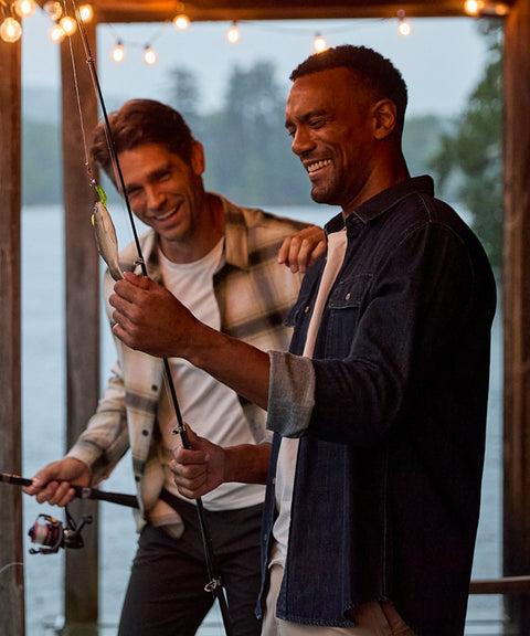 Two men fishing from a covered dock at dusk, one in a plaid overshirt and one in dark denim.