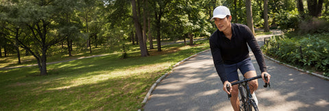 A man riding a bicycle wearing a black long-sleeve athletic shirt, dark shorts, and a white cap.