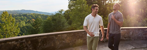 Two men walking and talking outdoors on a stone terrace with a lush green forest and hills in the background, during bright golden hour sunlight.