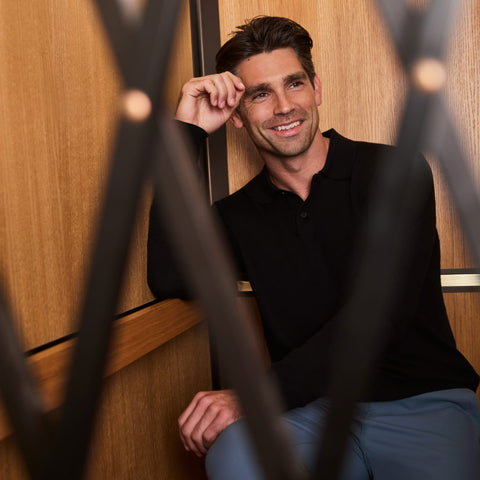 A black knit long-sleeve polo worn with blue trousers, seen through a dark lattice screen.