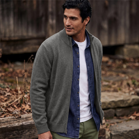 A man in a gray full-zip sweater over a denim shirt, standing against weathered wood siding.
