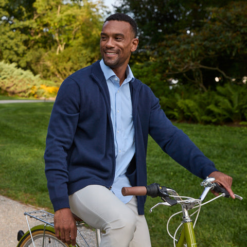 A cyclist in an open navy full-zip sweater over a light blue shirt, beside a bike in a park.