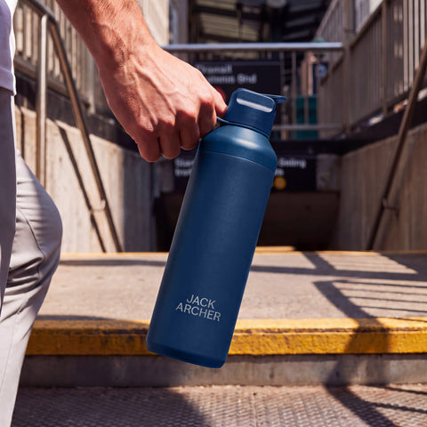 A navy Jack Archer insulated bottle carried by its lid loop at a transit station entrance.