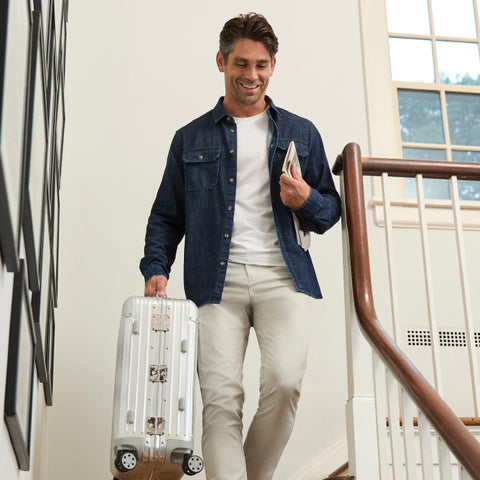 A traveler in an indigo denim shirt and stone chinos carrying a metal suitcase down a staircase.
