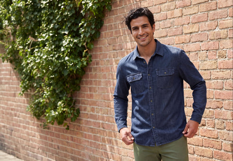 A man wearing a dark blue denim button-up shirt and olive green pants, standing against a brick wall with green ivy climbing up it.