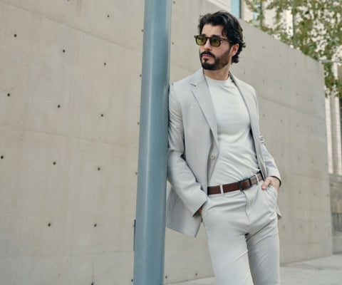Man in light gray blazer and matching pants with white tee and sunglasses leaning against a concrete pillar