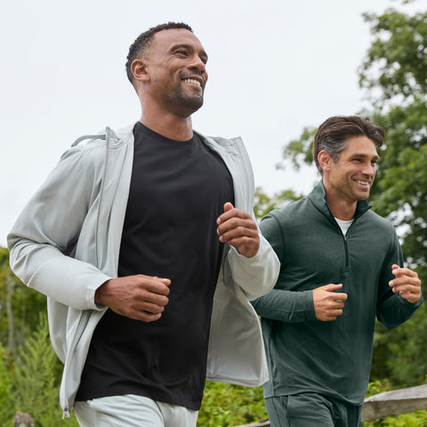 Two men jogging outdoors, one in an open gray hooded jacket over a black tee, one in a dark green quarter-zip.